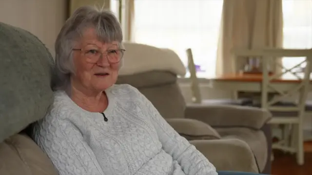 Wendy Martin, a pensioner sitting on the sofa in her home. She is wearing a grey Aran jumper.
