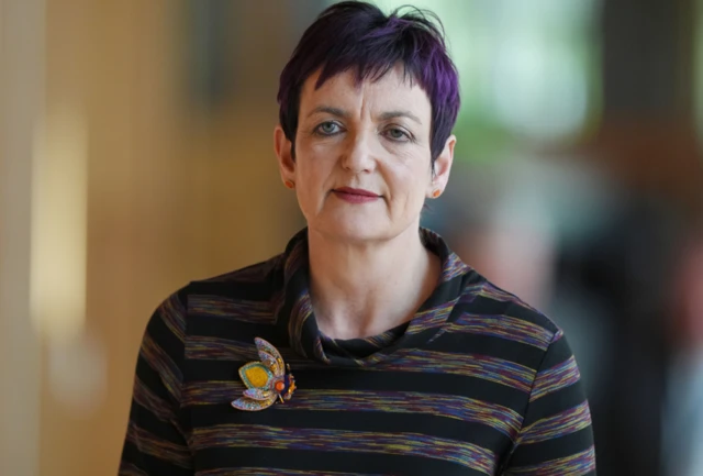 A tightly framed portrait shows a person facing the camera in an indoor setting with a softly blurred background. The person has short, dark hair and is wearing a striped, dark-coloured top. A distinctive butterfly-shaped brooch is pinned on the left side of the top, adding a bright, decorative detail. The background features warm tones and soft lighting, with indistinct shapes suggesting an interior corridor or room. The focus is sharp on the face and upper body, with the background intentionally out of focus to draw attention to the subject.