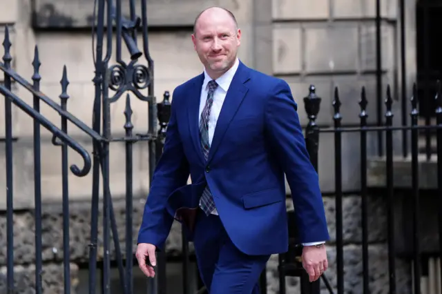 Neil Gray, in blue suit, walking.