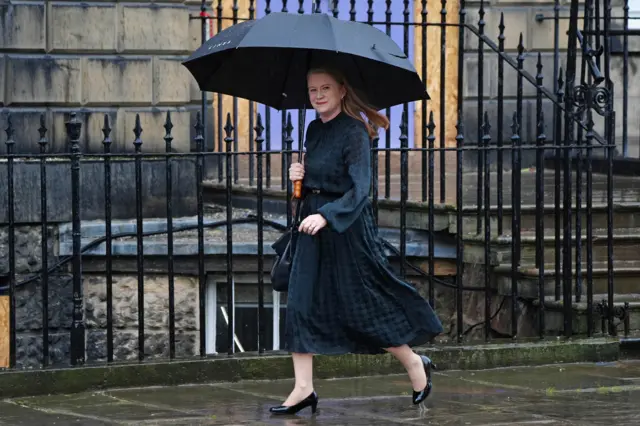 On a very rainy day and with brolly up, Shirley-Anne Somerville in a dark blue dress walks along the street and smiles