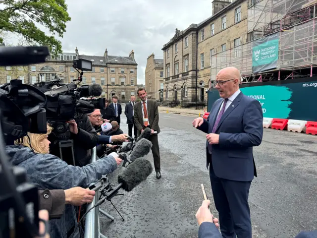 John Swinney in suit and purple tie speaking to lots of reporters outside Bute House