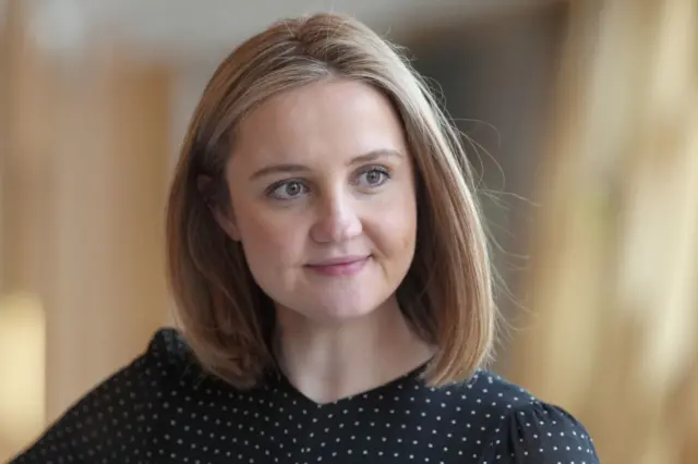 A tightly framed portrait shows a person facing the camera indoors, set against a softly blurred background. The person has short hair and is wearing a dark, striped top with a rounded neckline. A colourful butterfly-shaped brooch is pinned on the left side of the top, standing out against the darker fabric. The background features warm, muted tones with indistinct shapes, suggesting a corridor or interior public space. The lighting is soft and even, with the focus kept sharp on the face and upper body while the background remains out of focus.