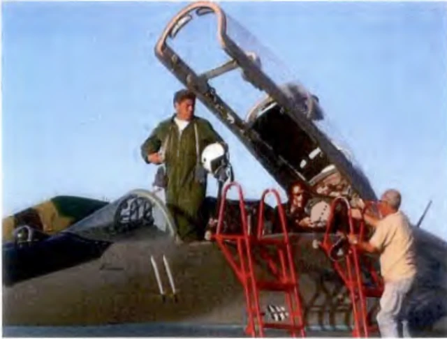 Luis Raul Gonzalez-Pardo Rodriguez is seen wearing a dark green flight suit and standing in the cockpit of a MiG fighter jet. Two red ladders and a pair of crew assist him.
