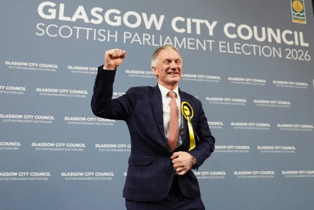 A man wearing an SNP rosette raises his fist and smiles at the Glasgow City Council Scottish Parliament Election 2026 count venue, standing in front of a blue branded backdrop.