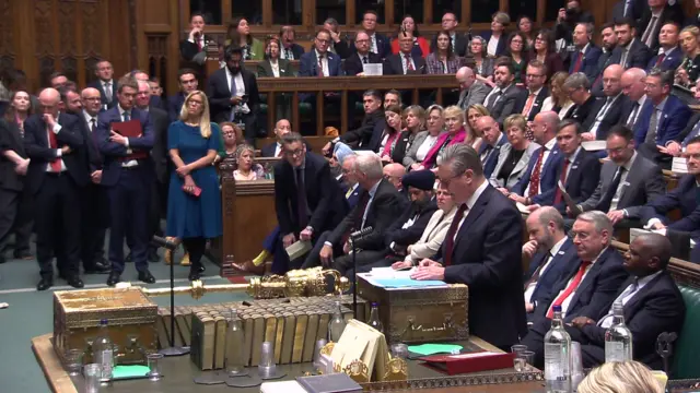 Starmer stands and speaks at the dispatch box. Labour MPs can be seen lining the benches behind him and a number can be seen standing at the far side of the House of Commons