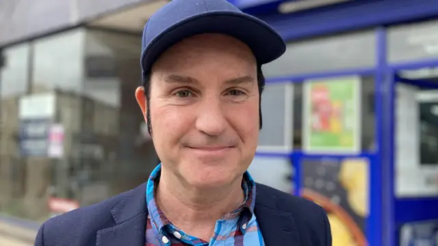 A man with a blue cap and blazer and a blue and red checked shirt.