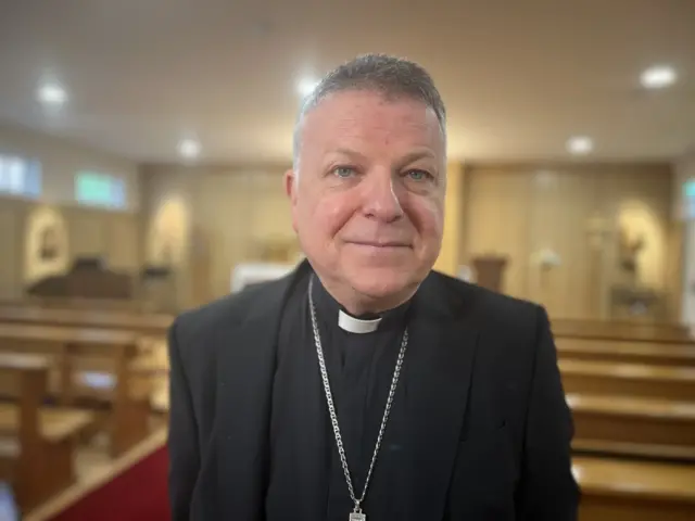 A priest in black suit, shirt and with dog collar and silver crucifix, smiles at the camera in a church