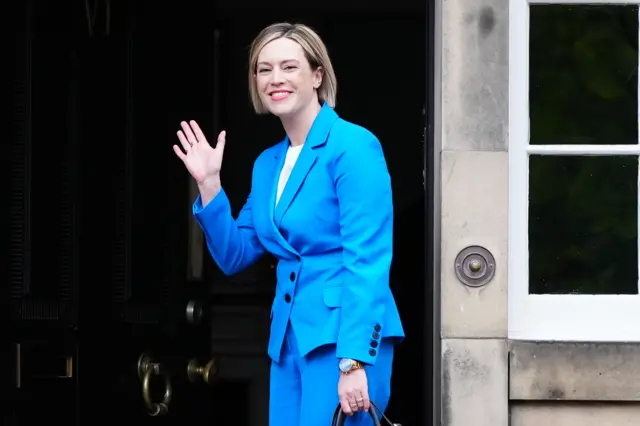 Jenny Gilruth in a blue suit waving as she walks into Bute House.