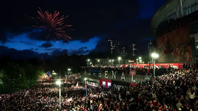Arsenal fans celebrate while fireworks go off outside the Emirates