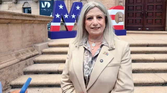 A woman stands in front of a building in Miami. She wears a beige suit jacket