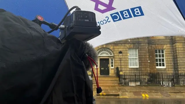 A TV camera under a BBC brolly, facing Bute House