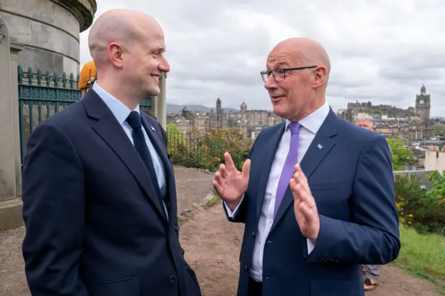 Stephen Flynn and John Swinney are two bald men in dark blue suits with light coloured shirts and blue and purple ties (respectively). They are talking and laughing on top of Calton Hill in Edinburgh. The Balmoral Hotel can be seen behind them.
