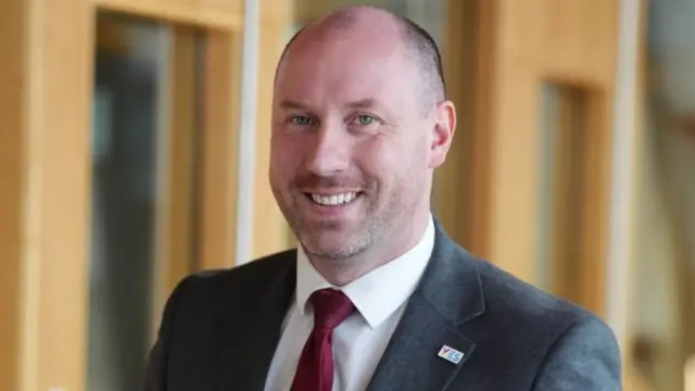 Neil Gray, who is bald and is wearing a suit with burgundy tie. He is smiling and has a YES badge on his lapel.