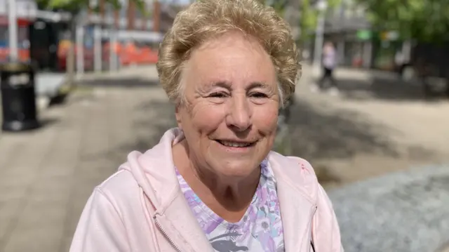 An elderly lady wearing a purple and while floral t-shirt and a pink hoodie.