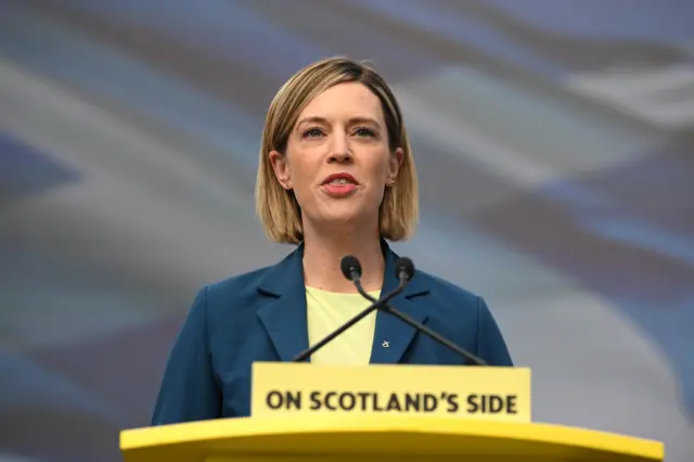 A person stands behind a lectern delivering a speech into two microphones positioned closely together. The person is wearing a dark blue blazer over a light-coloured top, with short, neatly styled hair. The lectern is bright yellow and features clearly visible text reading: “ON SCOTLAND’S SIDE”. The background is blurred, with soft grey and blue tones suggesting an outdoor or large public setting. The image is tightly framed around the upper body, focusing attention on the speaker and the podium.