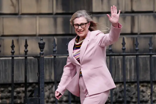 Gillian Martin is wearing a pink suit, stripey blouse, and tinted specs. She is waving and smiling broadly while walking.