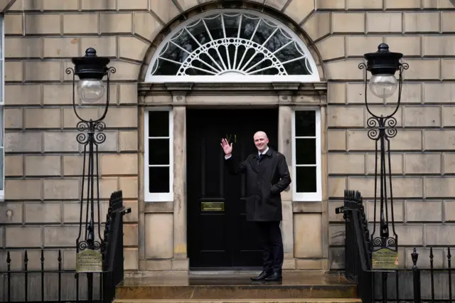 Stephen Flynn, in black suit, tie and coat with white shirt, smiles and waves outside Bute House