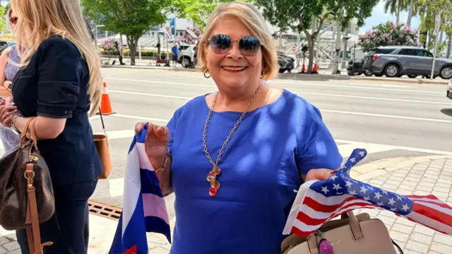 A woman stands outside in Miami. She wears a blue shirt and holds an American and a Cuban flag