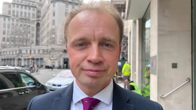 A close up image of a middle aged man with thinning blonde hair in a suit with a purple tie.