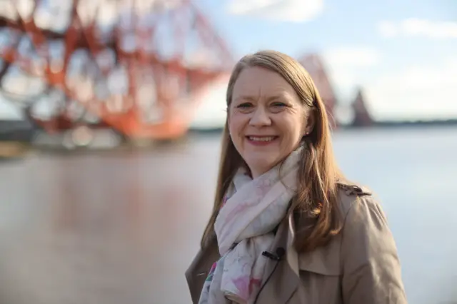 Somerville in scarf and coat standing in front of the Forth Bridge on a sunny day