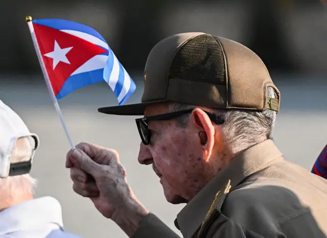 Raul Castro in a cap and dark glasses waves a small Cuban flag