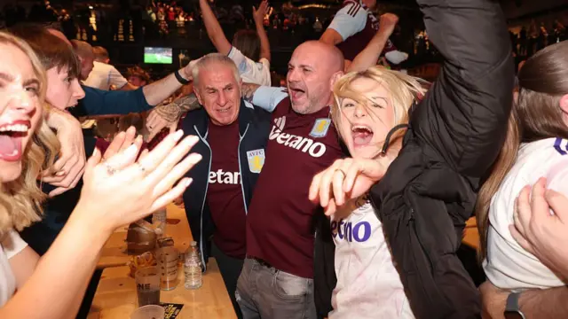 Two women at a table with two other men celebrate. They are wearing Villa tops and raise their fists in the air