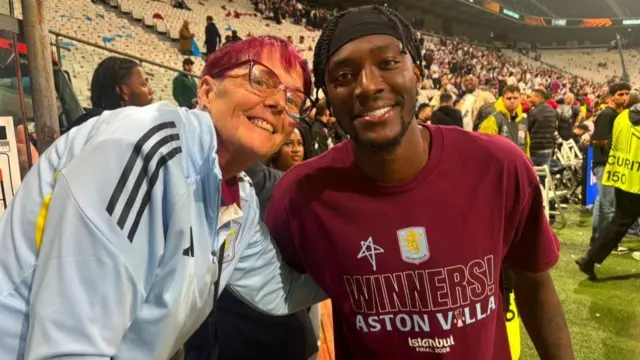 Jane Bissell, a woman with short pink hair and a blue jacket, smiles as she poses for a photo with an Aston Villa player in a stadium.