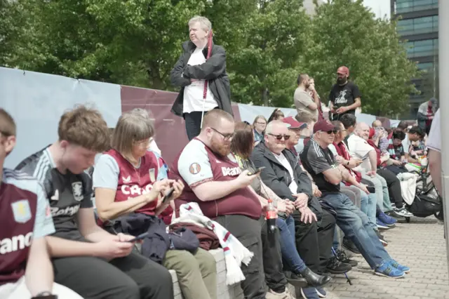 A line of Aston Villa fans, most wearing Villa shirts, sat down on a small wall on the side of a footpath.