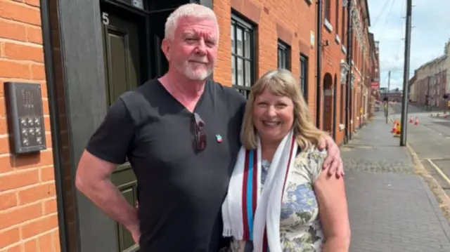 A man with short white hair and wearing a black shirt has his arm around the shoulder of a woman with long blonde hair, a flowery top and a white, claret and blue scarf. They are standing on the side of a road.