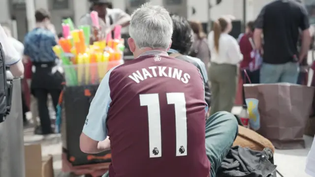 A man with grey hair sits down with his back to us. He is wearing a claret and blue Aston Villa shirt which says "Watkins 11" on the back.