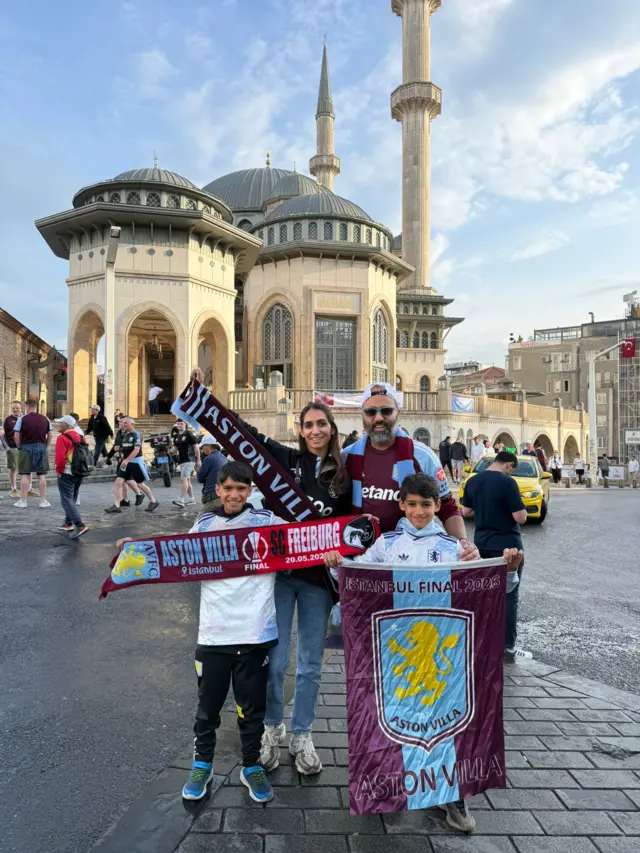 Ravi and Anaeka stand in an Istanbul street with sons Arjun and Abhi holding Aston villa flags and scarves