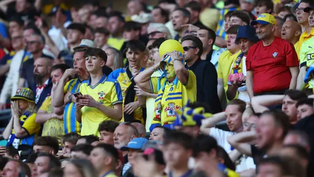 Saints fans in the stands in yellow and blue with their hands on their heads