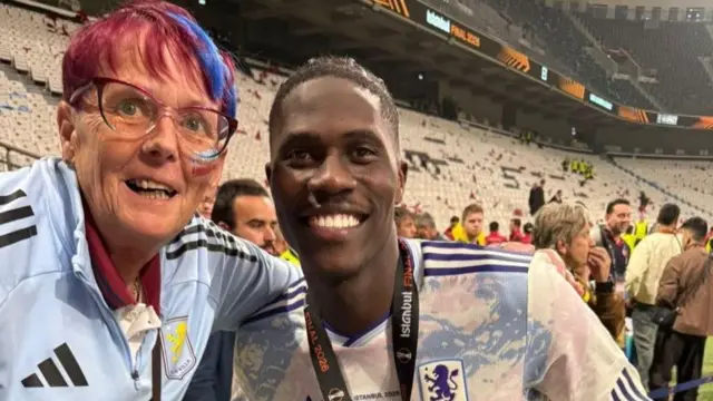 Jane Bissell, a woman with short pink hair and a blue jacket, smiles as she poses for a photo with an Aston Villa player in a stadium.