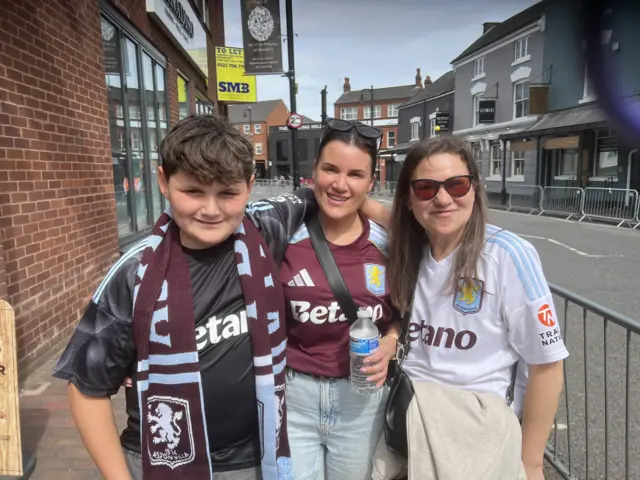Charlie, Becky and Vivian stand in the street wearing Villa tops and Charlie has a scarf around his neck