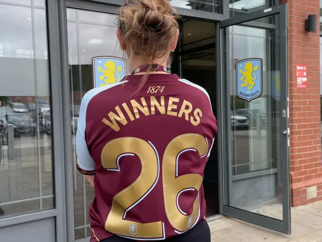 A woman with her back to the camera, wearing an Aston Villa shirt in claret and blue that says Winners 26