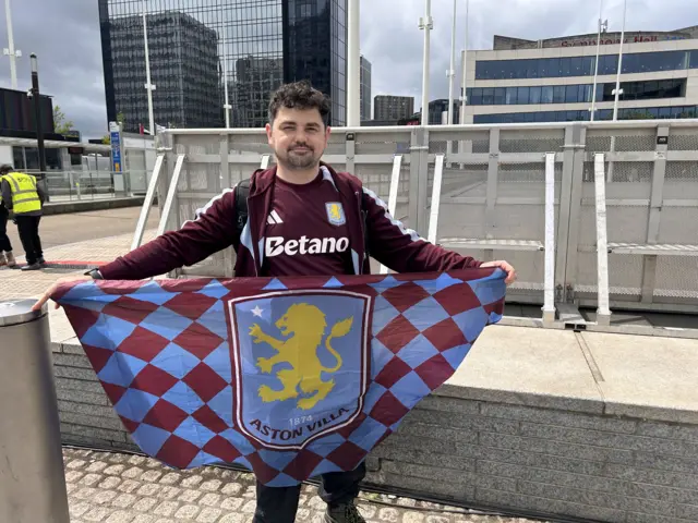 Daryl stands in an Aston Villa top, and holding up a club flag in front of him