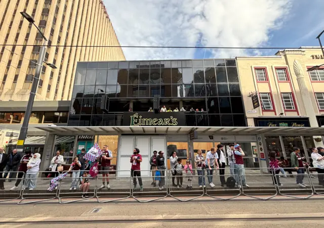 People line up at the side of the road behind a barrier, outside Eimear's Irish bar in Birmingham. The bar is closed for work but builders can be seen watching on the balcony.