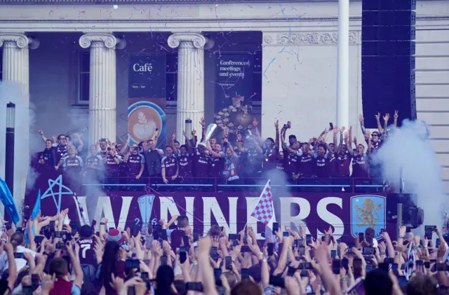 Villa players on a bus lift the trophy in front of fans.