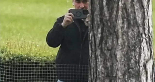 A young man holding a camera phone, standing behind a tree