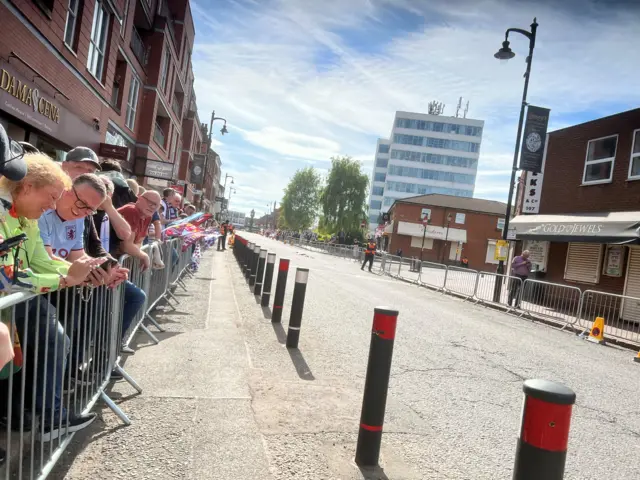 People lined up along a street behind a barrier as they await the Aston Villa parade bus. The other side of the street, where a barrier has also been erected, is emtpy.