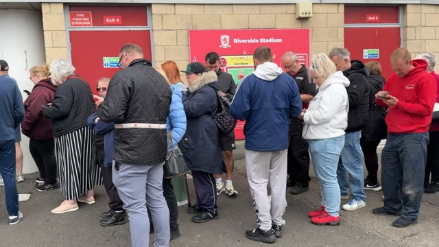 About two dozen people standing in a line outside the stadium. They are mainly looking at their phones. There are red doors behind them and a red sign which reads: "Riverside Stadium."