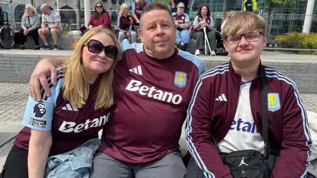 A woman and two men wearing Aston Villa outfits sit huddled together in a square in Birmingham city centre.