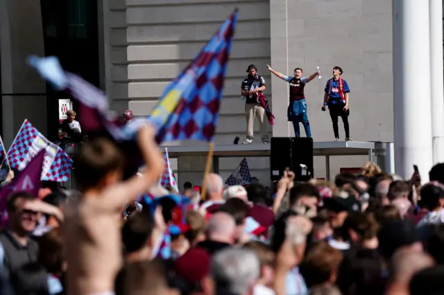 A crowd of people, some holding up Aston Villa flags, gathered in Birmingham city centre. Three men are standing on a bus shelter in the background cheering.