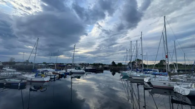 Thick fluffy clouds across the sky over a marina full of sail boats. There is a small bit of blue sky breaking through the cloud