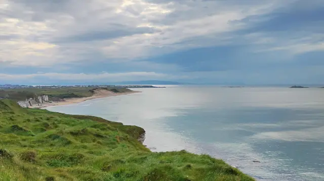 A view from the cliffs above a beach and the sea. The sky is cloudy but blue and spots of sun come through