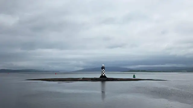 A lighthouse in the middle of a big water way. It's grey outside in the water and the skies, which have heavy cloud