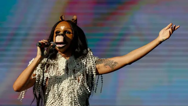 Woman wearing a horse mask over her head singing into microphone with one hand in the air