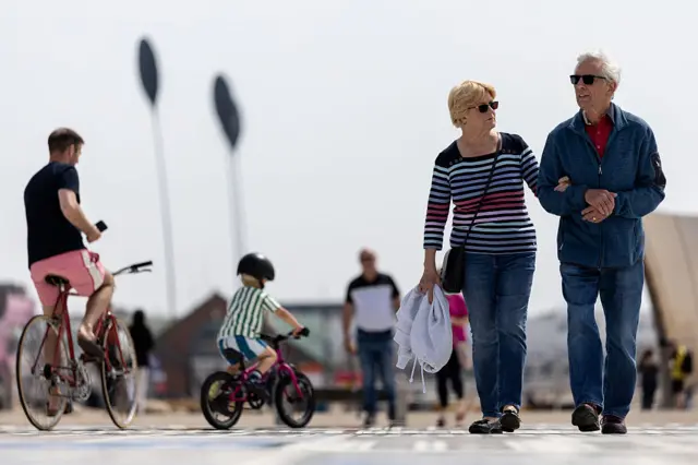 An elderly couple wearing sunglasses make their way Holidaymakers make their way along Blackpool Promenade as a child and middle aged man cycle past them