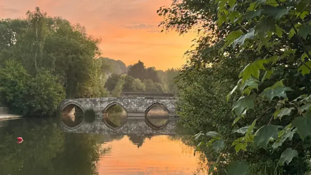 A sun rising causing the sky to be orange. There's an old stone bridge over a wide river surrounded by trees