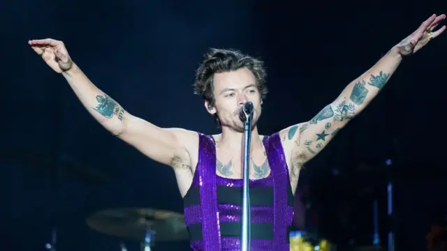 Harry Styles in a purple and black suit singing into a microphone with his hands in the air